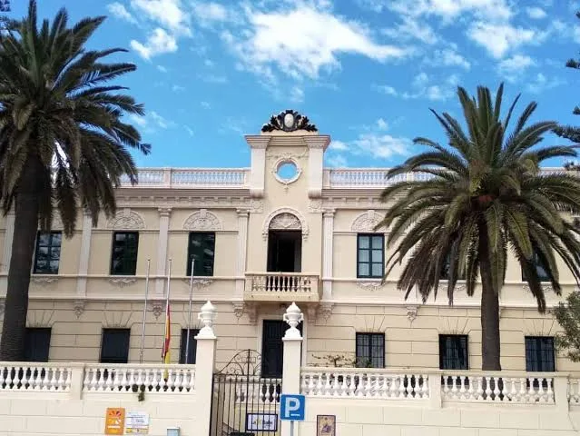 Edificio de arquitectura neoclásica de color crema con palmeras, balconada blanca y reloj en la fachada principal.