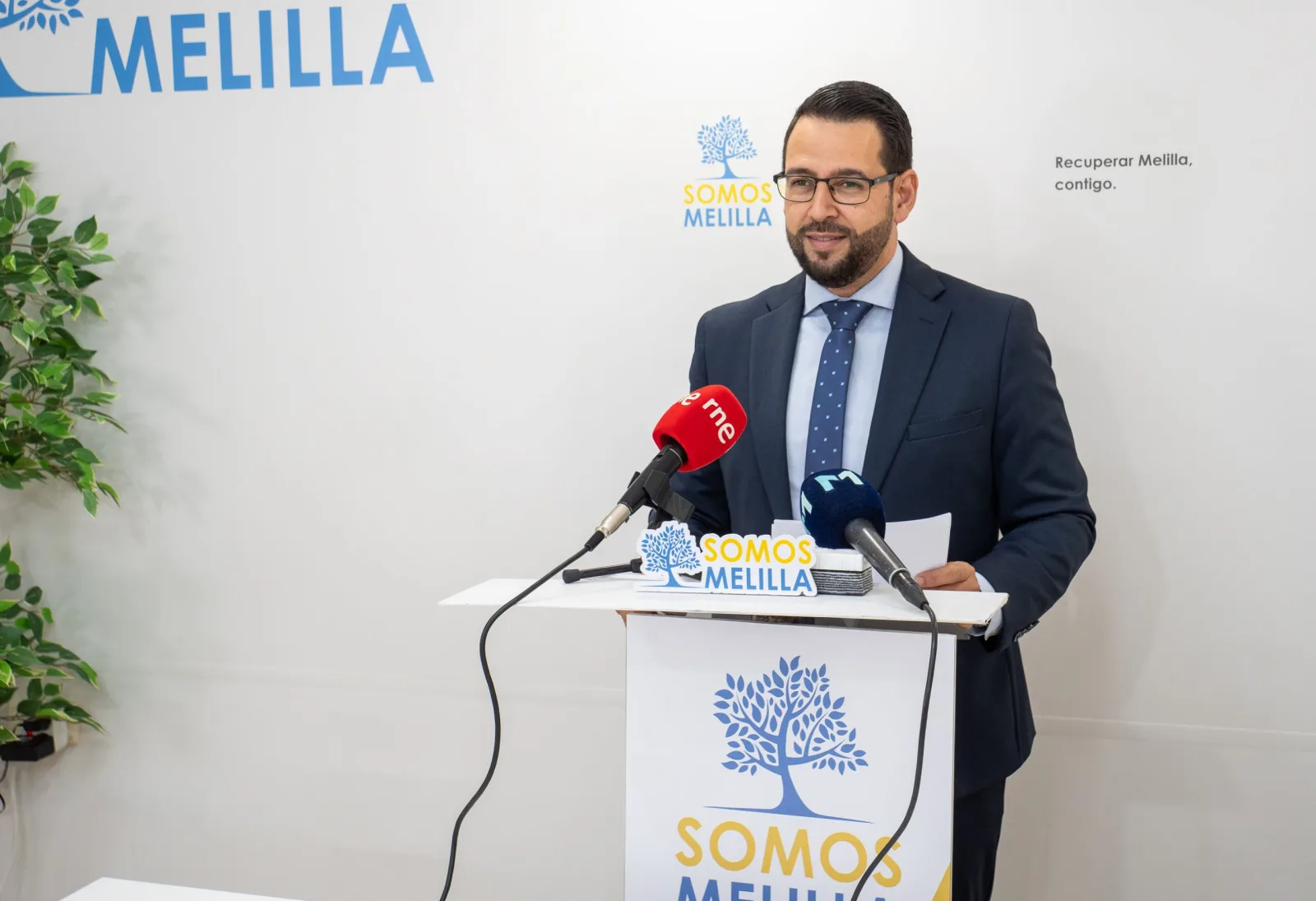 Hombre de traje oscuro y gafas ante un podio con micrófono en una sala con logos de Somos Melilla.