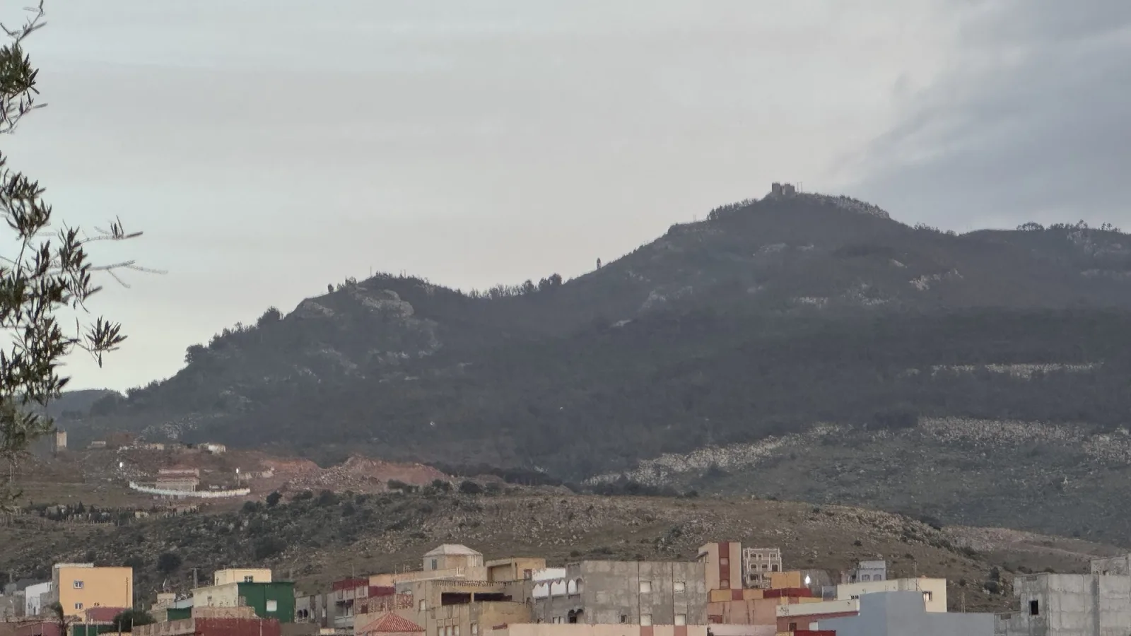 El monte Gurugú visto desde la zona de Melilla, con su cumbre coronada por las ruinas de una fortificación, laderas de matorral y casas en primer plano bajo un cielo nublado.