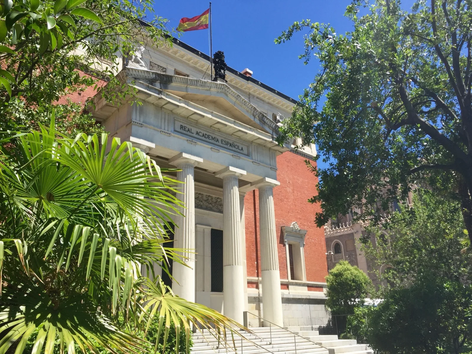 Fachada neoclásica con columnas de la Real Academia Española, enmarcada por palmeras y árboles, con la bandera en lo alto.