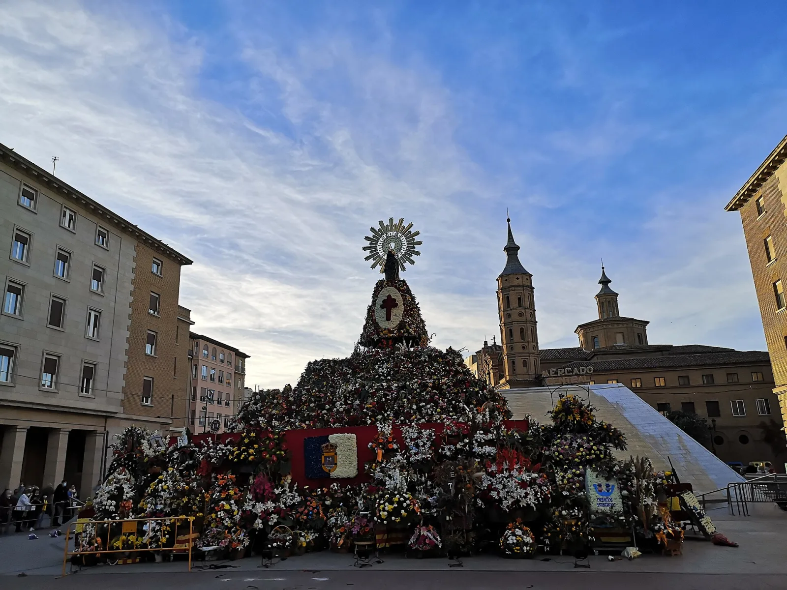 Gran manto de flores con la imagen de la Virgen del Pilar en lo alto, en una plaza de Zaragoza rodeada de edificios, al atardecer.
