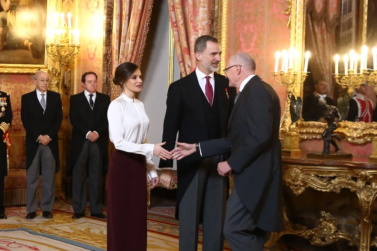 Don Felipe y doña Letizia saludan a un invitado en un salón del Palacio Real, con candelabros encendidos y cortinajes al fondo.