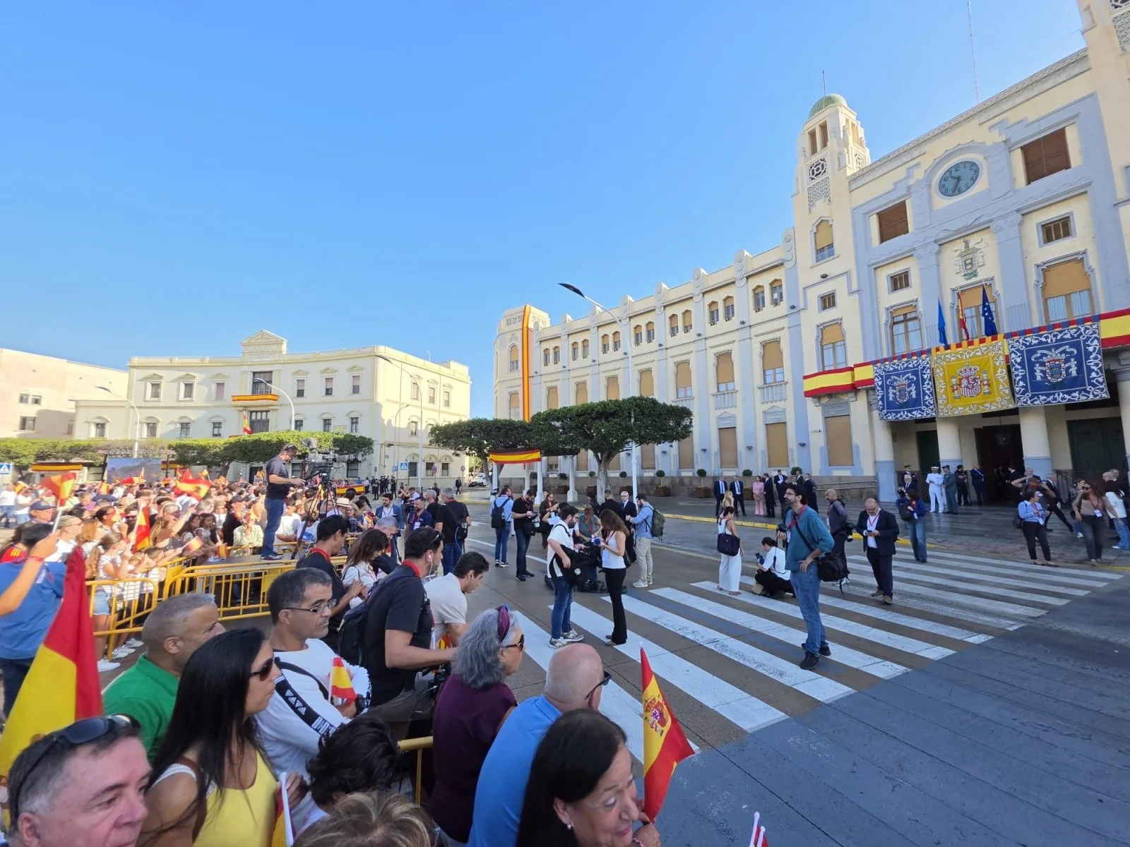 Vecinos esperando a los Reyes en la plaza de España de Melilla, con banderas y el palacio de la Asamblea al fondo.