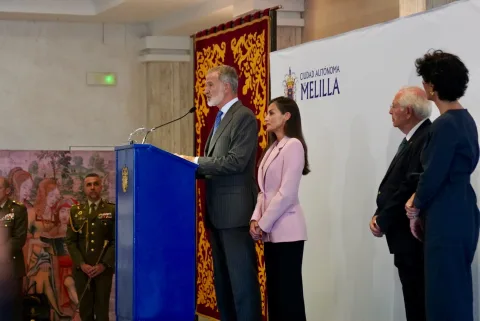 El Rey Felipe VI habla en el atril junto a la Reina Letizia ante el panel de la Ciudad Autónoma de Melilla, con militares al fondo.