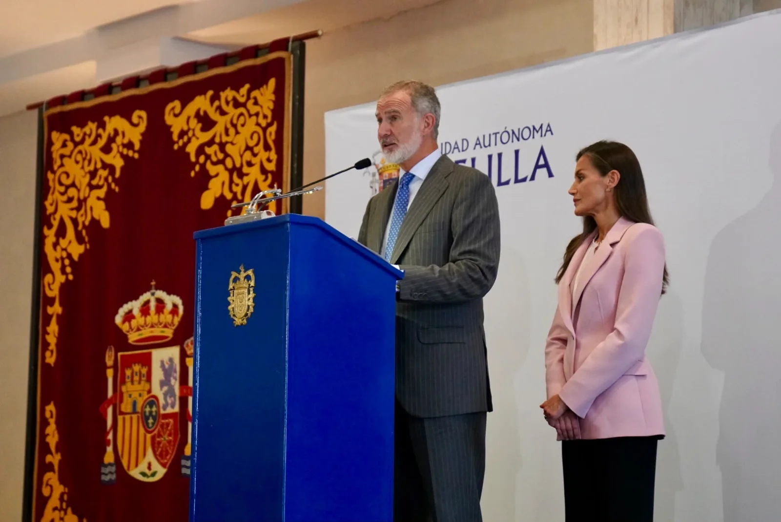 El Rey Felipe VI pronuncia su discurso en un atril azul junto a la Reina Letizia, con el escudo de España en un tapiz al fondo.