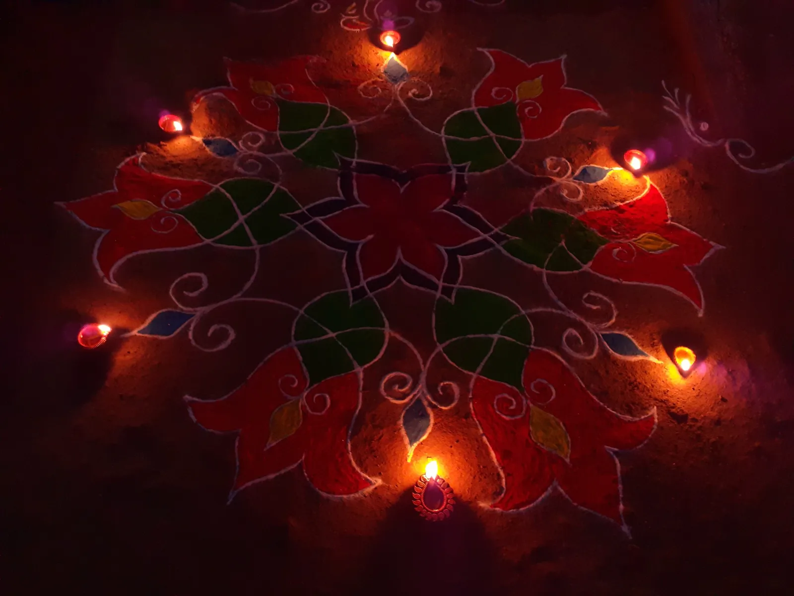 Rangoli de flores rojas y hojas verdes dibujado con polvos de colores en el suelo, rodeado de pequeñas lámparas de barro encendidas en la noche del Diwali