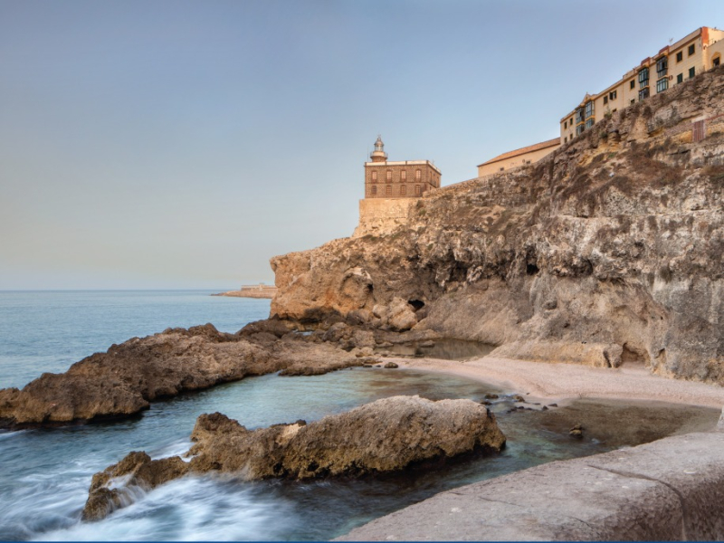 Costa rocosa con una pequeña cala, rocas en el agua y edificios sobre un acantilado al fondo.
