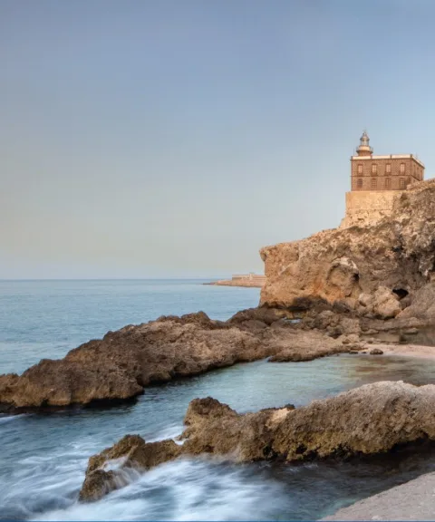 Una edificación histórica con cúpula sobre un acantilado rocoso junto al mar, con rocas y agua en primer plano.