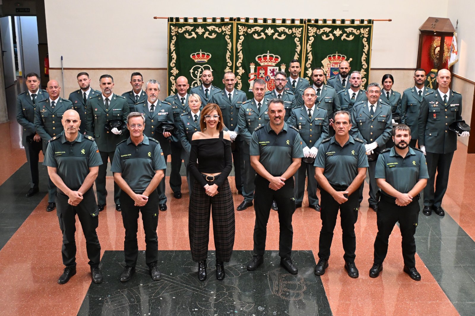 Un grupo de personas uniformadas posa en filas ante unos tapices verdes con escudos, con una mujer de negro en el centro.