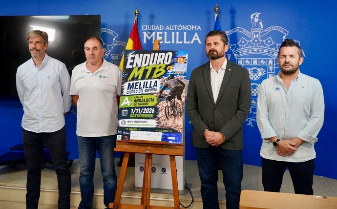 Cuatro hombres de pie junto a un cartel de Enduro MTB sobre un caballete, ante un fondo azul con el escudo de Melilla.