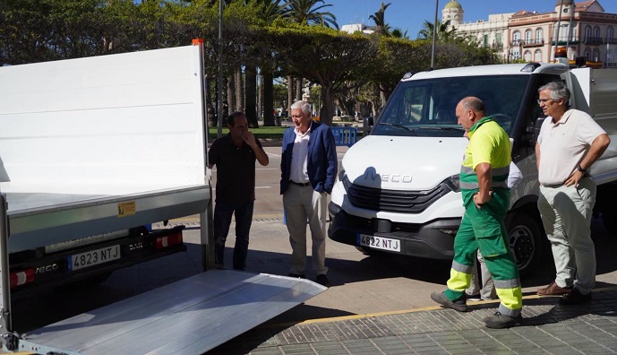 Cuatro hombres junto a un camión volquete blanco con la plataforma trasera bajada, en una vía urbana con palmeras.