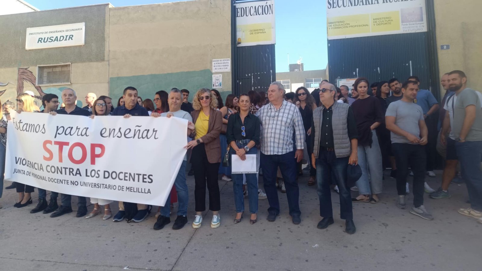 Un grupo de personas sostiene una pancarta grande que dice Estamos para enseñar STOP VIOLENCIA CONTRA LOS DOCENTES ante un centro educativo.