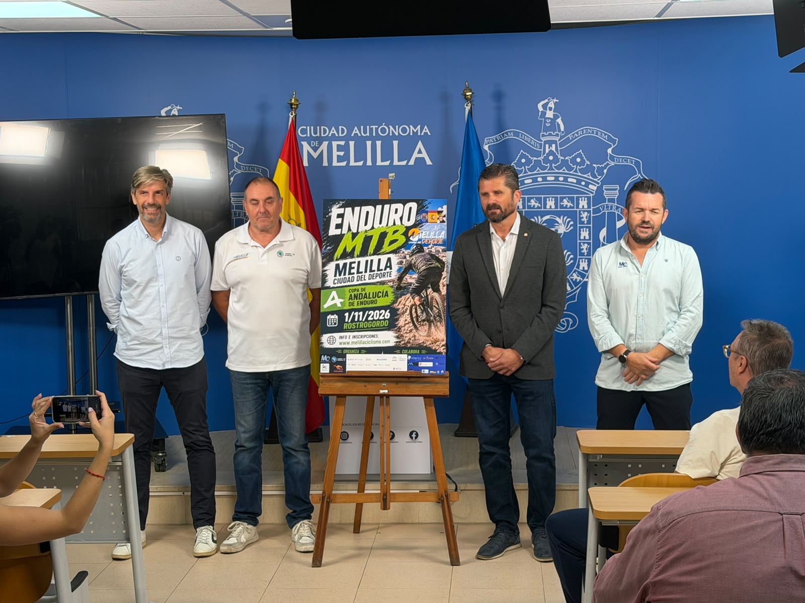 Cuatro hombres de pie junto a un cartel de Enduro MTB sobre un caballete, ante una pared azul con el escudo de Melilla.