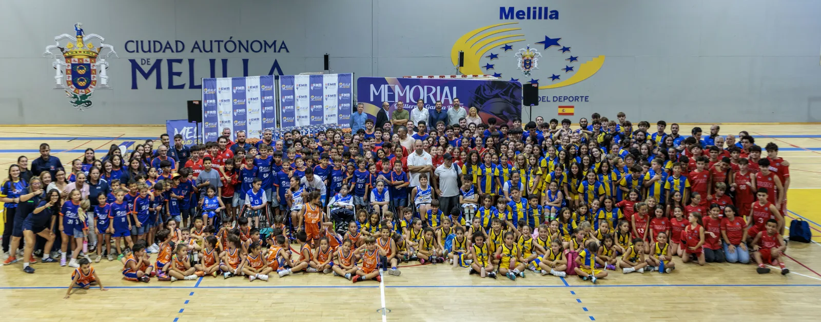 Gran grupo de niños y adultos, muchos con equipaciones deportivas, posando en una cancha cubierta ante carteles del Memorial.