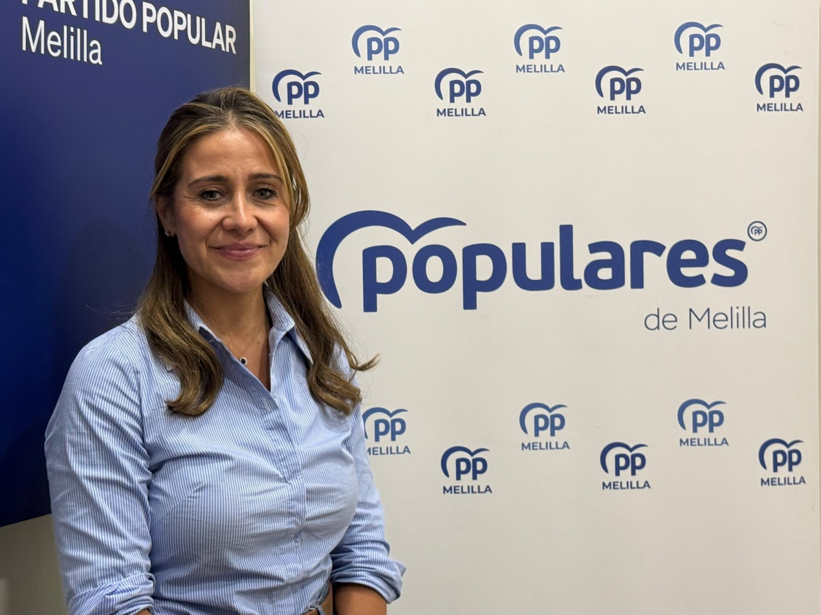 Una mujer con camisa azul de rayas posa sonriendo ante un panel con el logotipo del PP de Melilla.