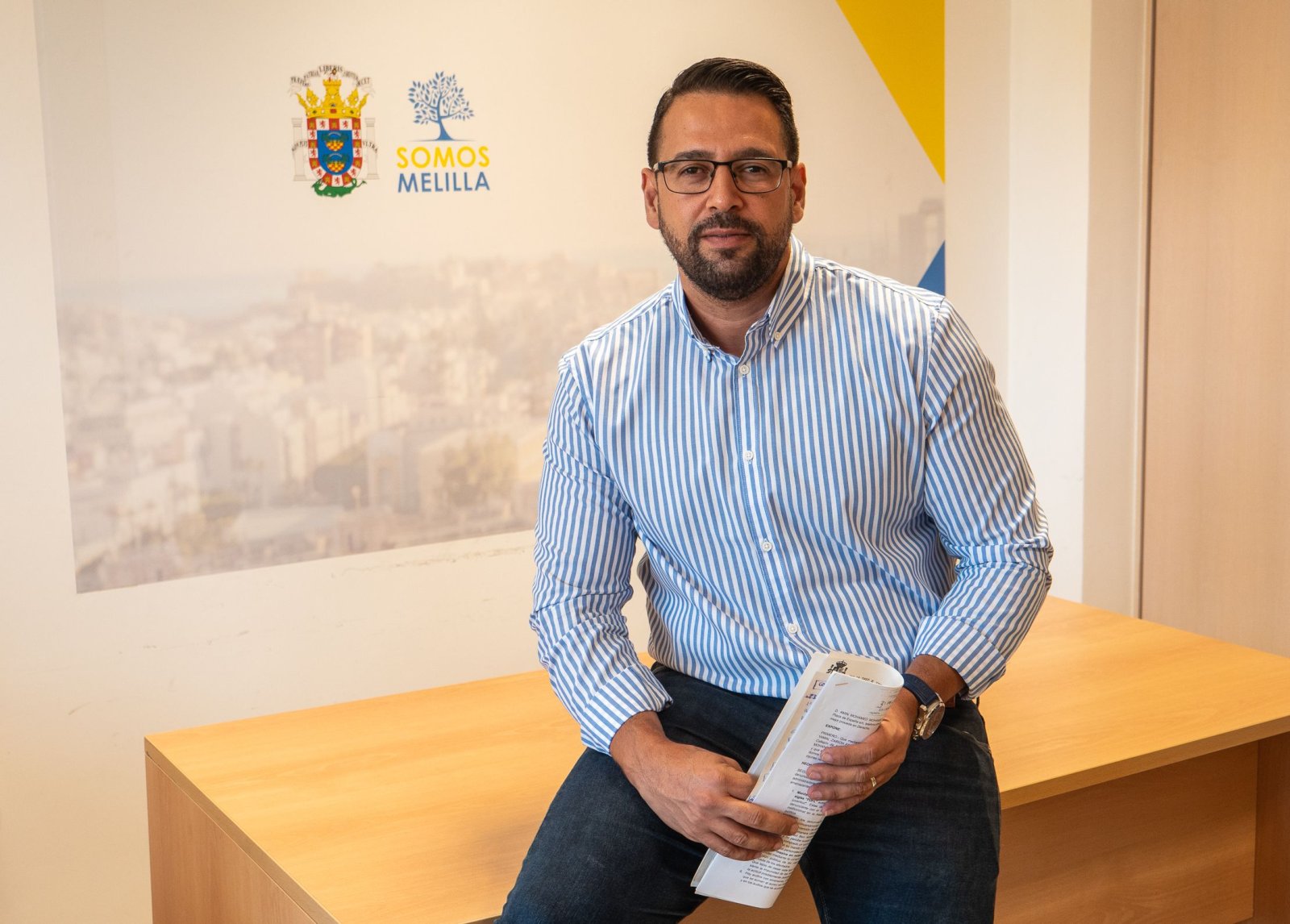 Un hombre con gafas y camisa de rayas azules sentado sobre una mesa de madera, con un folleto en las manos y un cartel de Somos Melilla detrás.