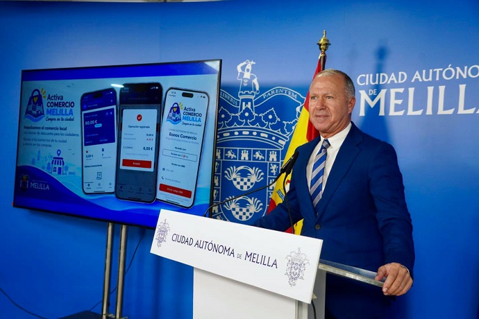 Un hombre trajeado habla ante un atril con el escudo de Melilla, junto a una pantalla que muestra una app de bonos.