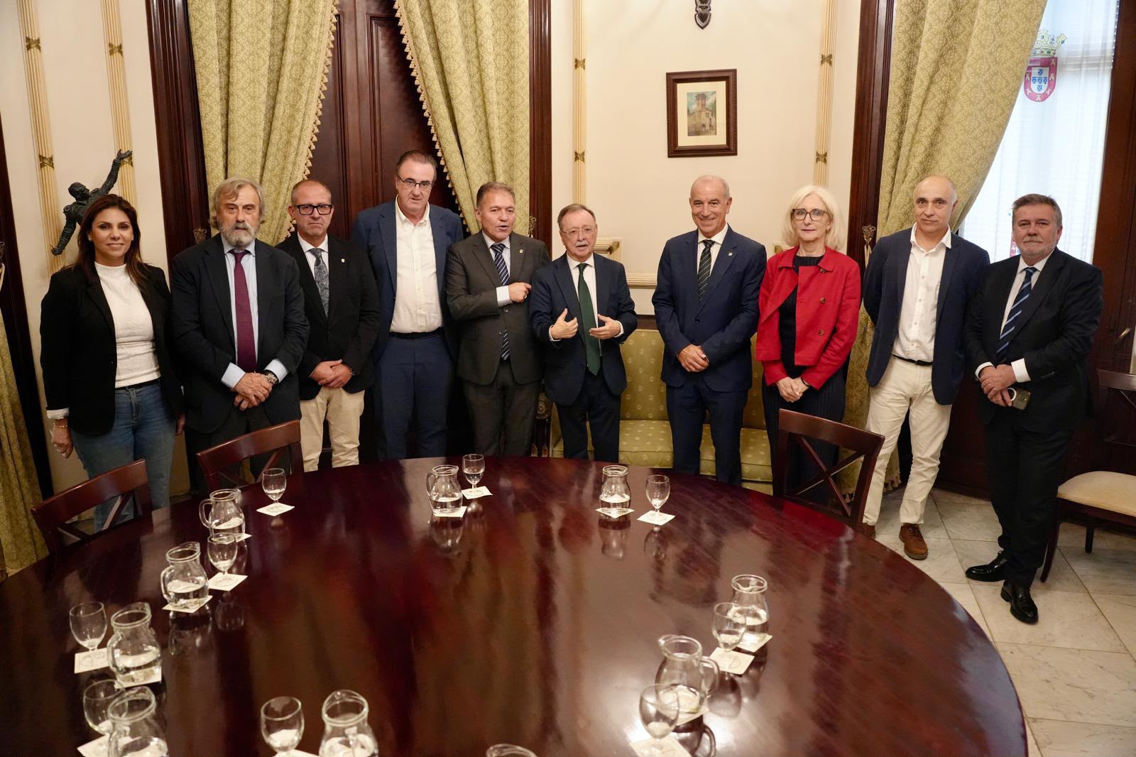 Diez personas trajeadas posan de pie tras una gran mesa ovalada de madera con jarras y vasos, en una sala con cortinas y cuadro.