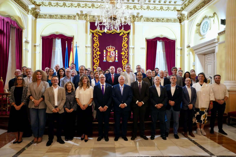 Un grupo numeroso de personas posa de pie en una sala noble con cortinajes rojos, banderas y un escudo bordado.