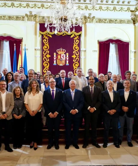 Un grupo numeroso de personas trajeadas posa de pie en una sala con cortinajes rojos, escudo y lámpara de araña.