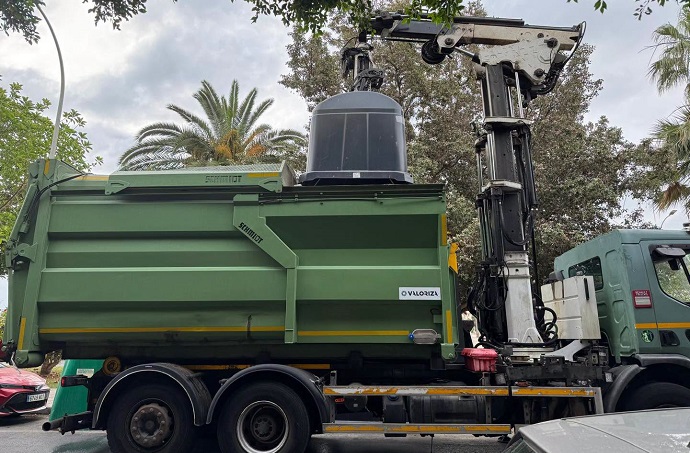 Camión de basura verde con grúa cargando un contenedor, junto a palmeras y otros vehículos.