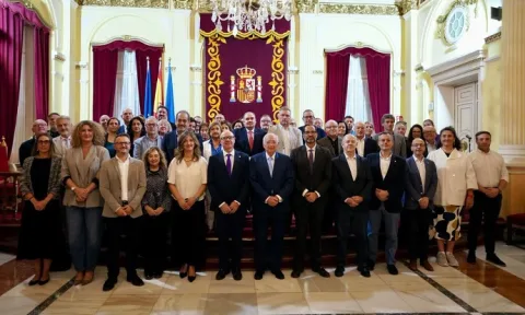 Un grupo numeroso de personas posa de pie en filas dentro de un salón con cortinajes rojos, banderas y un escudo en la pared.
