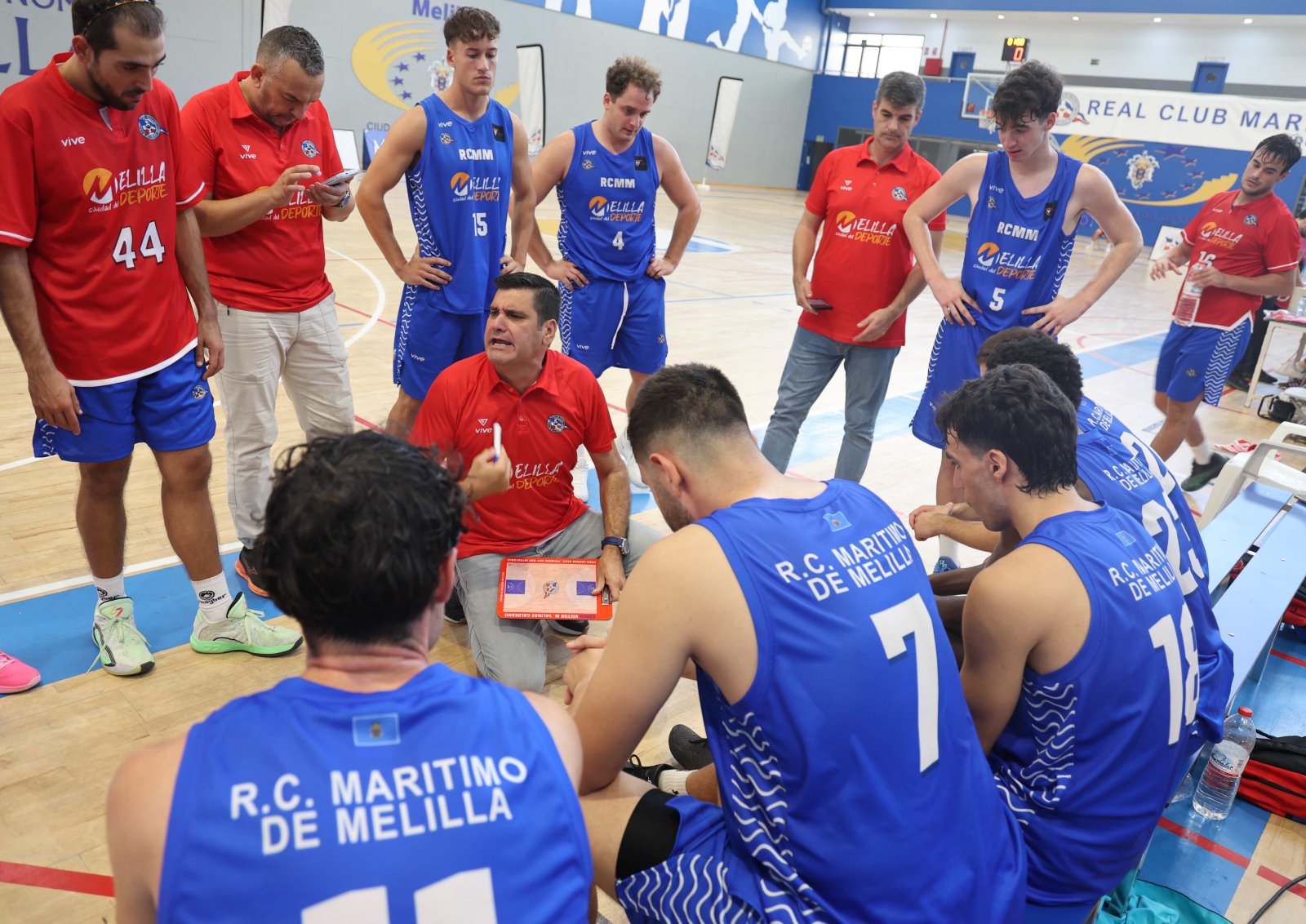 Jugadores de baloncesto con camisetas azules y rojas reunidos alrededor de un entrenador sentado en la pista.