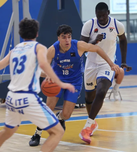 Un jugador de baloncesto con camiseta azul número 18 bota el balón mientras dos rivales de blanco lo defienden en una cancha cubierta.