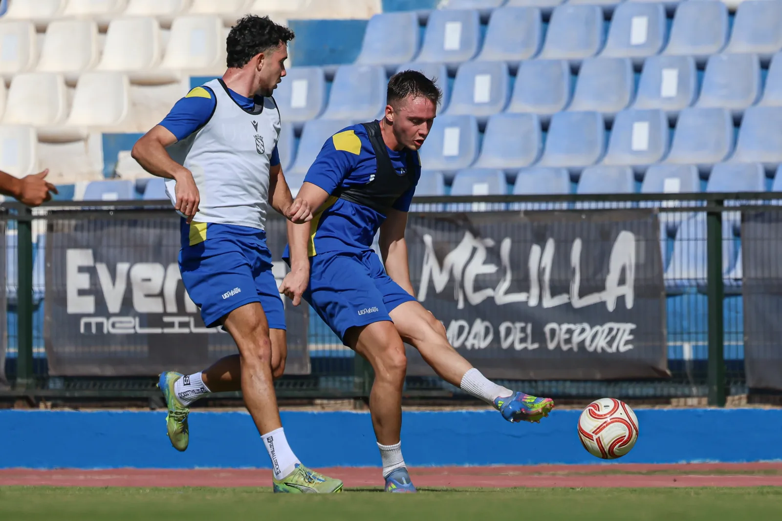 Dos futbolistas con equipación azul y blanca entrenan con un balón junto a una valla con pancartas y gradas azules al fondo.