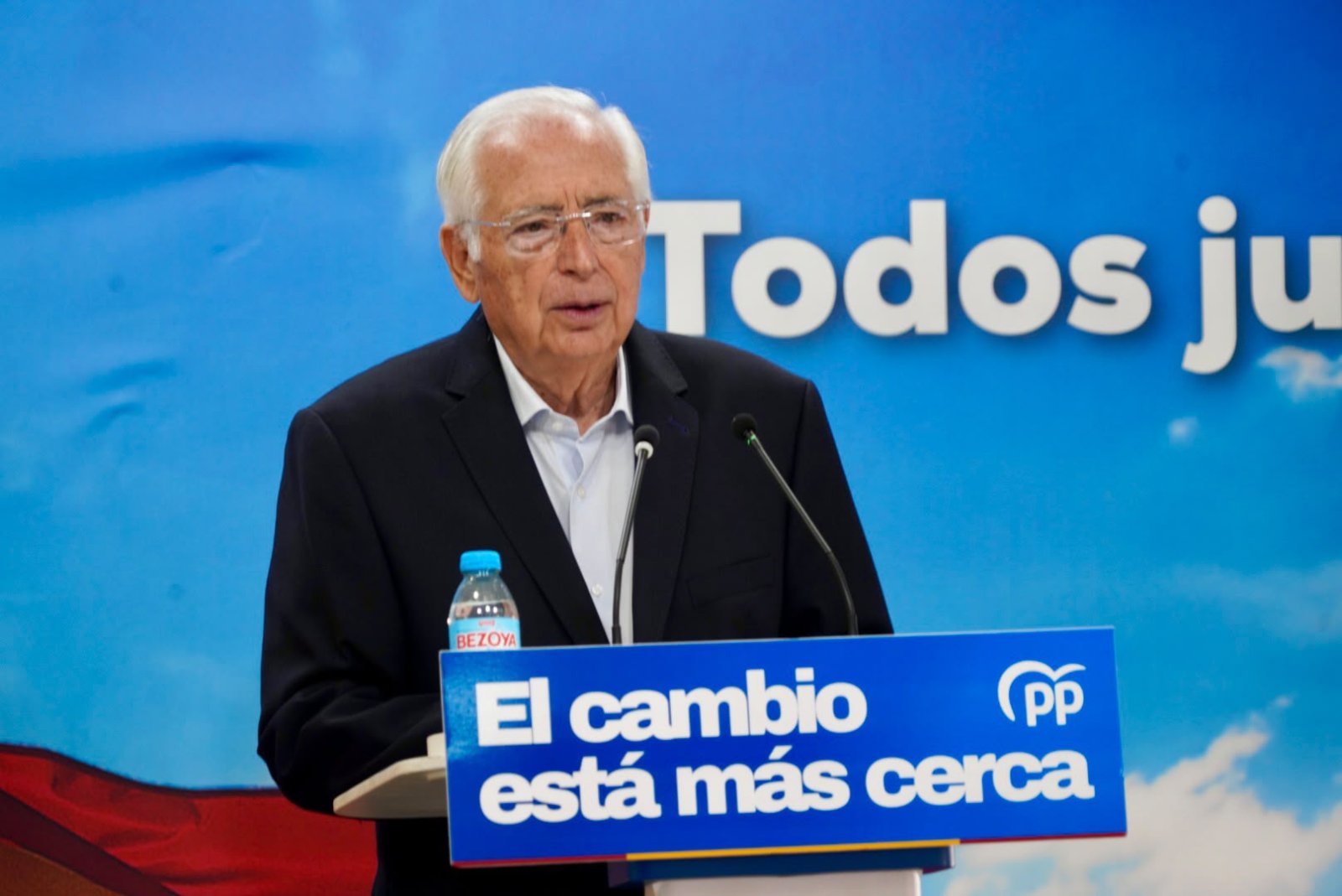 Un hombre mayor con gafas y traje oscuro habla ante dos micrófonos tras un atril azul con el lema El cambio está más cerca y el logo del PP.