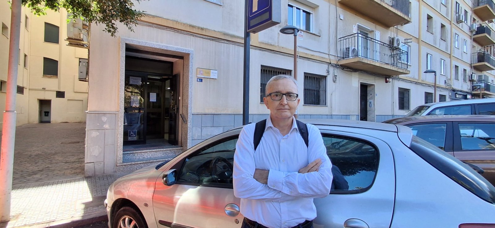 Un hombre mayor con gafas y camisa blanca, con los brazos cruzados, de pie junto a un coche plateado en una calle con edificios.