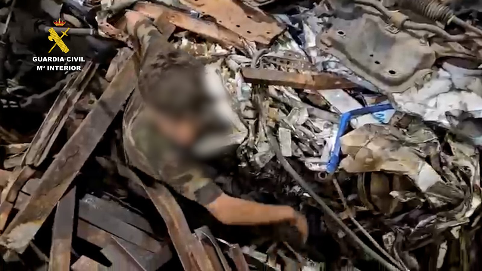 Una persona con ropa de camuflaje y el rostro difuminado entre chatarra y piezas metálicas, con el emblema de la Guardia Civil arriba a la izquierda.
