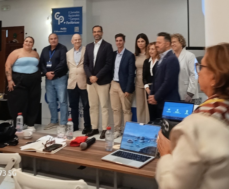 Un grupo de personas posa de pie tras una mesa con portátiles, botellas y un micrófono, en una sala con un cartel azul al fondo.
