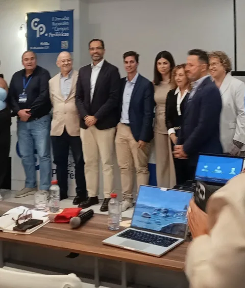 Un grupo de personas posa de pie tras una mesa con portátiles, botellas y un micrófono, ante un cartel de unas jornadas.