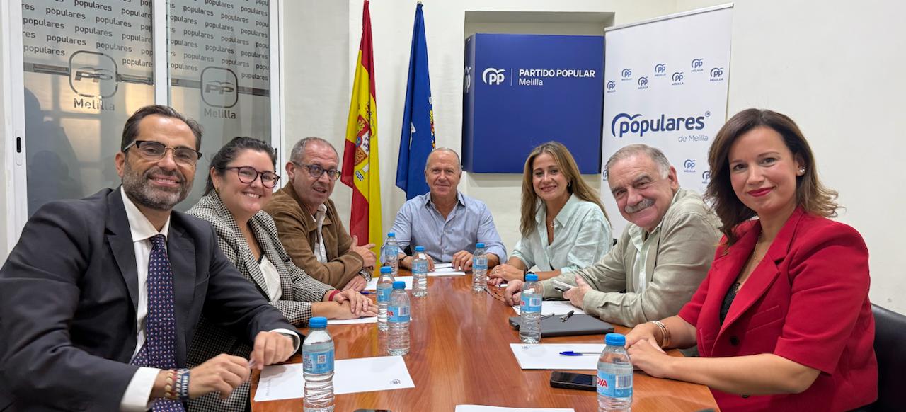 Siete personas sentadas alrededor de una mesa de reuniones con botellas de agua, banderas de España y la UE y un cartel del Partido Popular de Melilla al fondo.