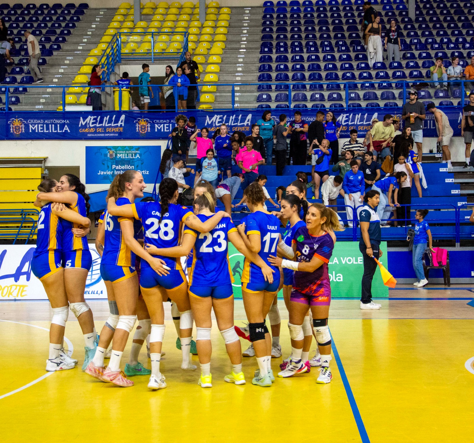 Jugadoras de voleibol abrazadas en la pista amarilla de un pabellón, con público en las gradas azules y amarillas.
