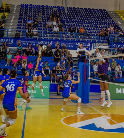 Jugadoras de voleibol en pleno partido junto a la red, con una rematando, ante gradas azules con público.