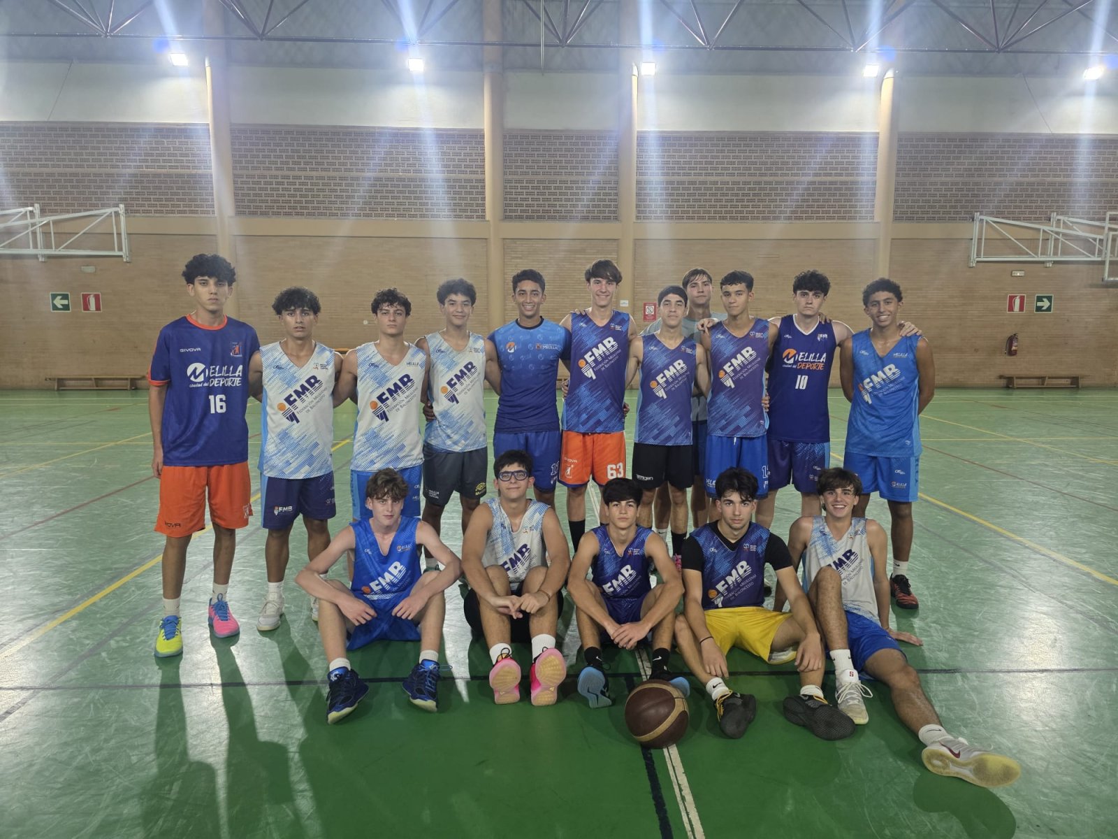 Un grupo de jugadores jóvenes de baloncesto posa en fila sobre una cancha cubierta, con un balón en el suelo.