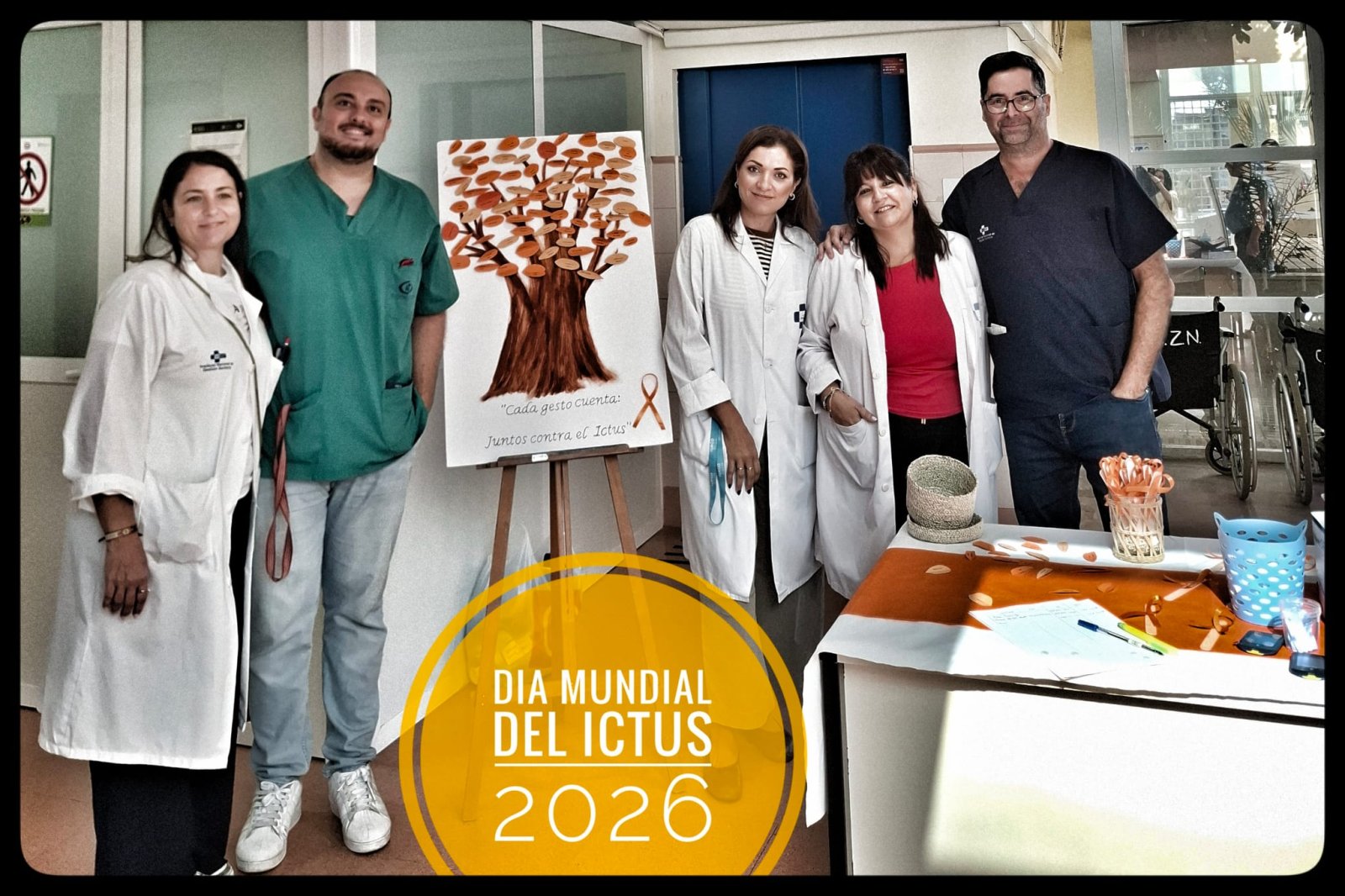Cinco personas con batas y pijama sanitario posan junto a un cartel con un árbol y una mesa informativa en un pasillo.