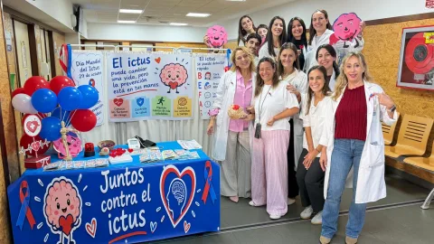 Un grupo de personas con batas blancas posa sonriendo junto a una mesa azul con carteles y globos sobre el ictus.