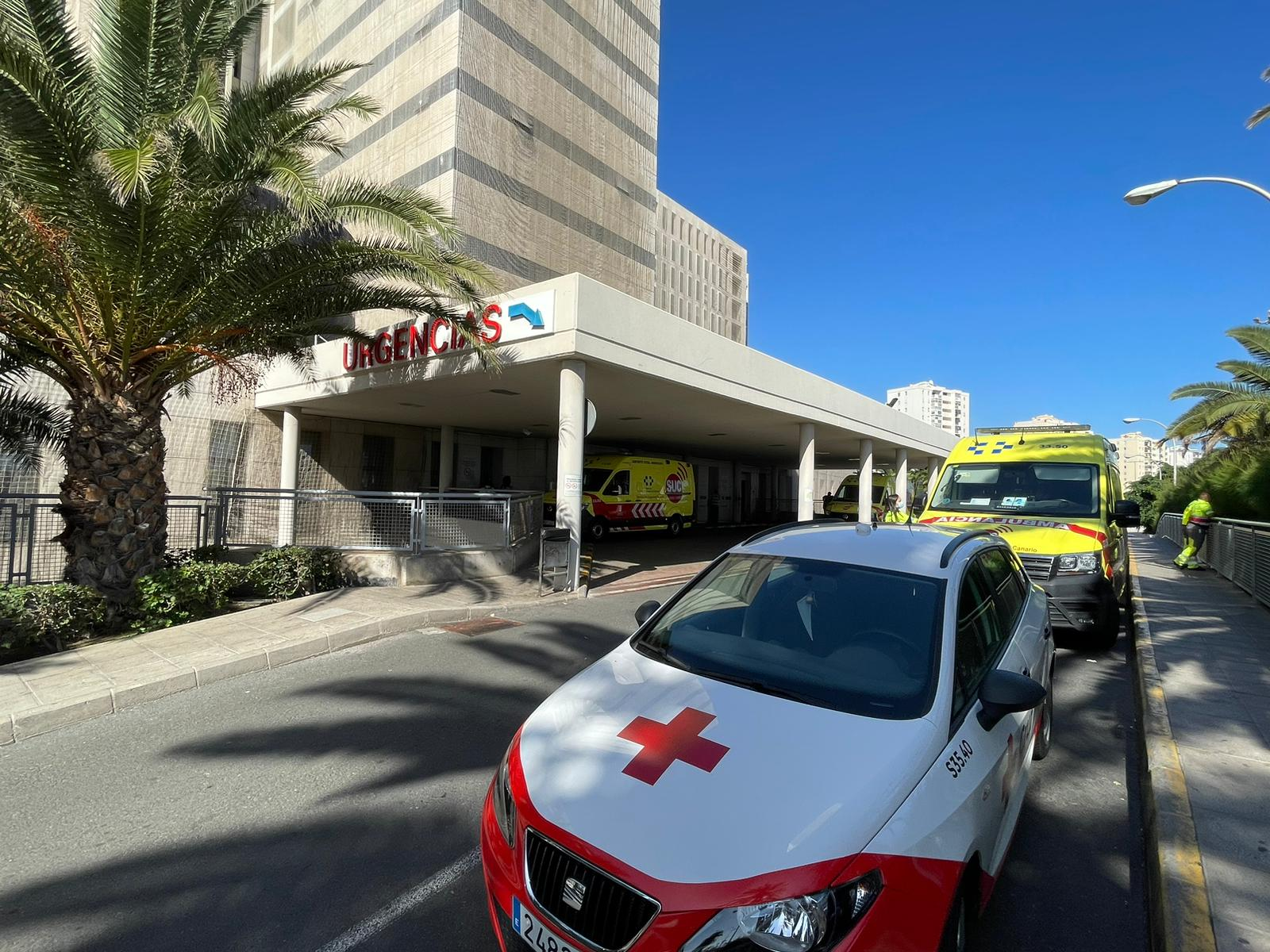 Ambulancias junto a la entrada de Urgencias de un hospital, con un coche blanco y rojo en primer plano.