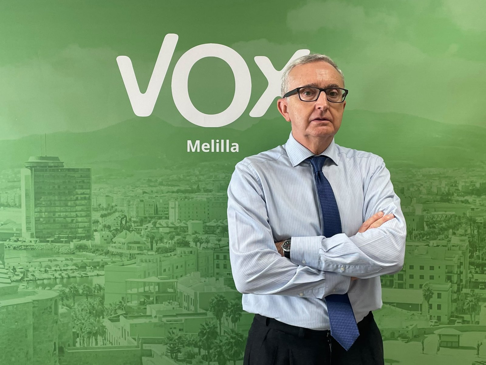 Hombre con gafas, camisa clara y corbata azul, con los brazos cruzados, ante un fondo verde con el logotipo de VOX Melilla y una vista urbana.
