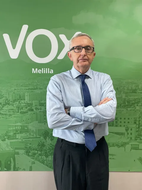 Hombre con gafas, camisa clara y corbata azul, con los brazos cruzados, ante un panel verde con el logotipo de Vox y la palabra Melilla.