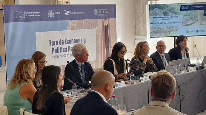 Varias personas sentadas tras una mesa larga en un foro, con un cartel de fondo y una pantalla encendida.