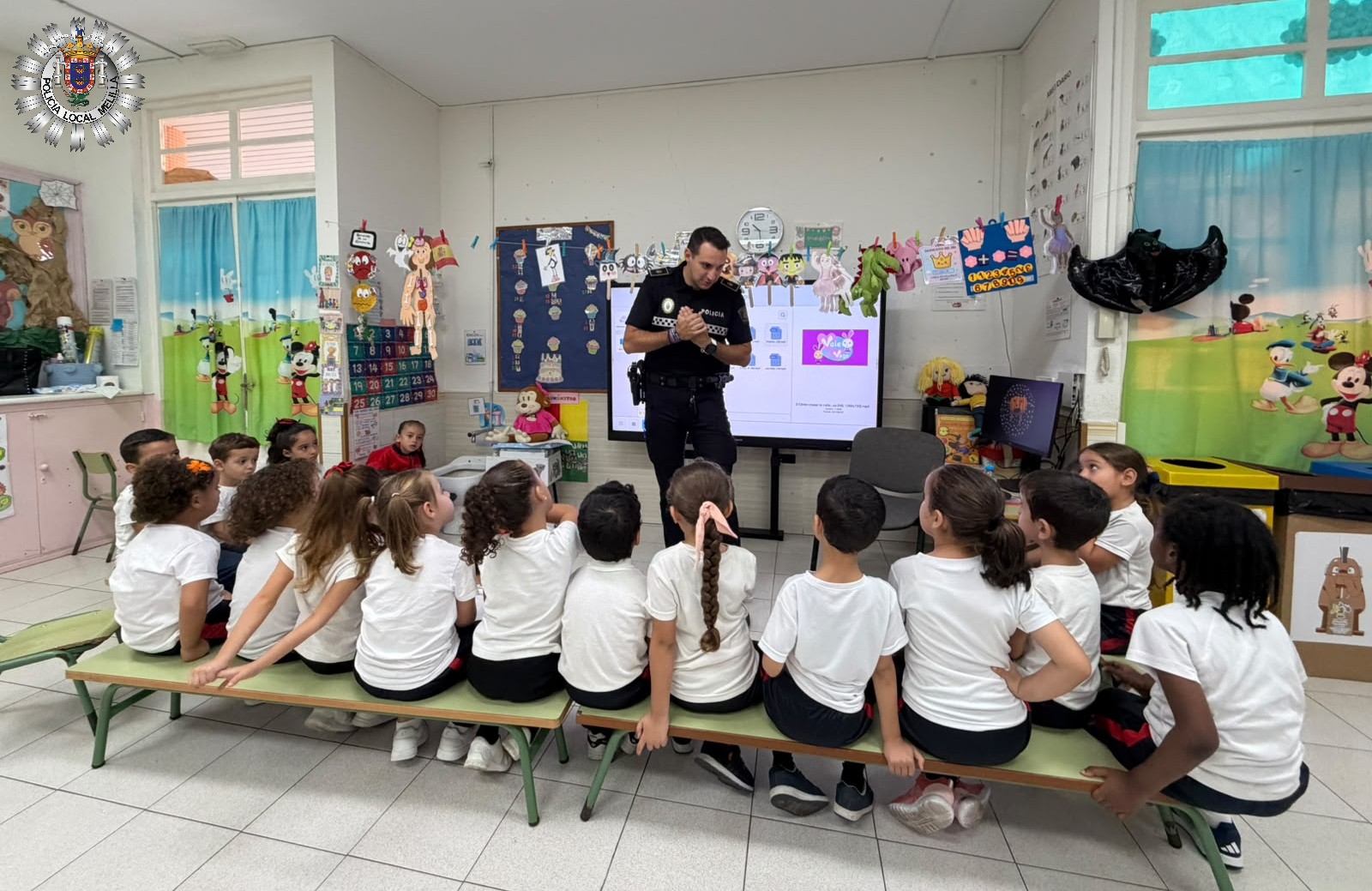 Un agente uniformado habla ante un grupo de niños sentados en bancos dentro de un aula con pantalla y dibujos.