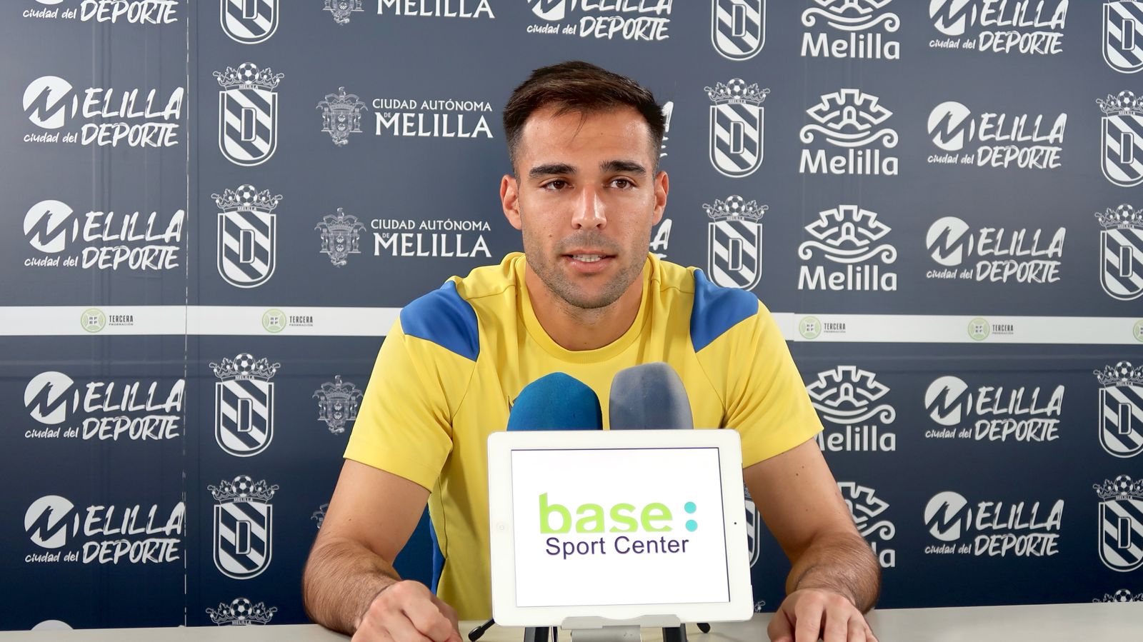Un hombre joven con camiseta amarilla y azul habla ante micrófonos y una tableta con el rótulo base: Sport Center, sobre un fondo con logotipos de Melilla.