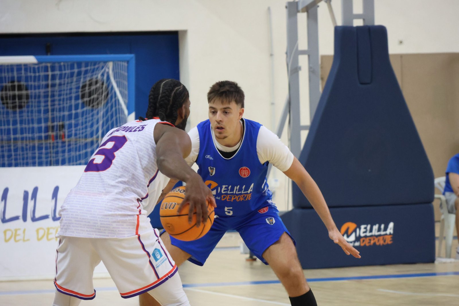 Dos jugadores de baloncesto, uno de espaldas con camiseta blanca y otro de frente con camiseta azul, en una cancha cubierta.