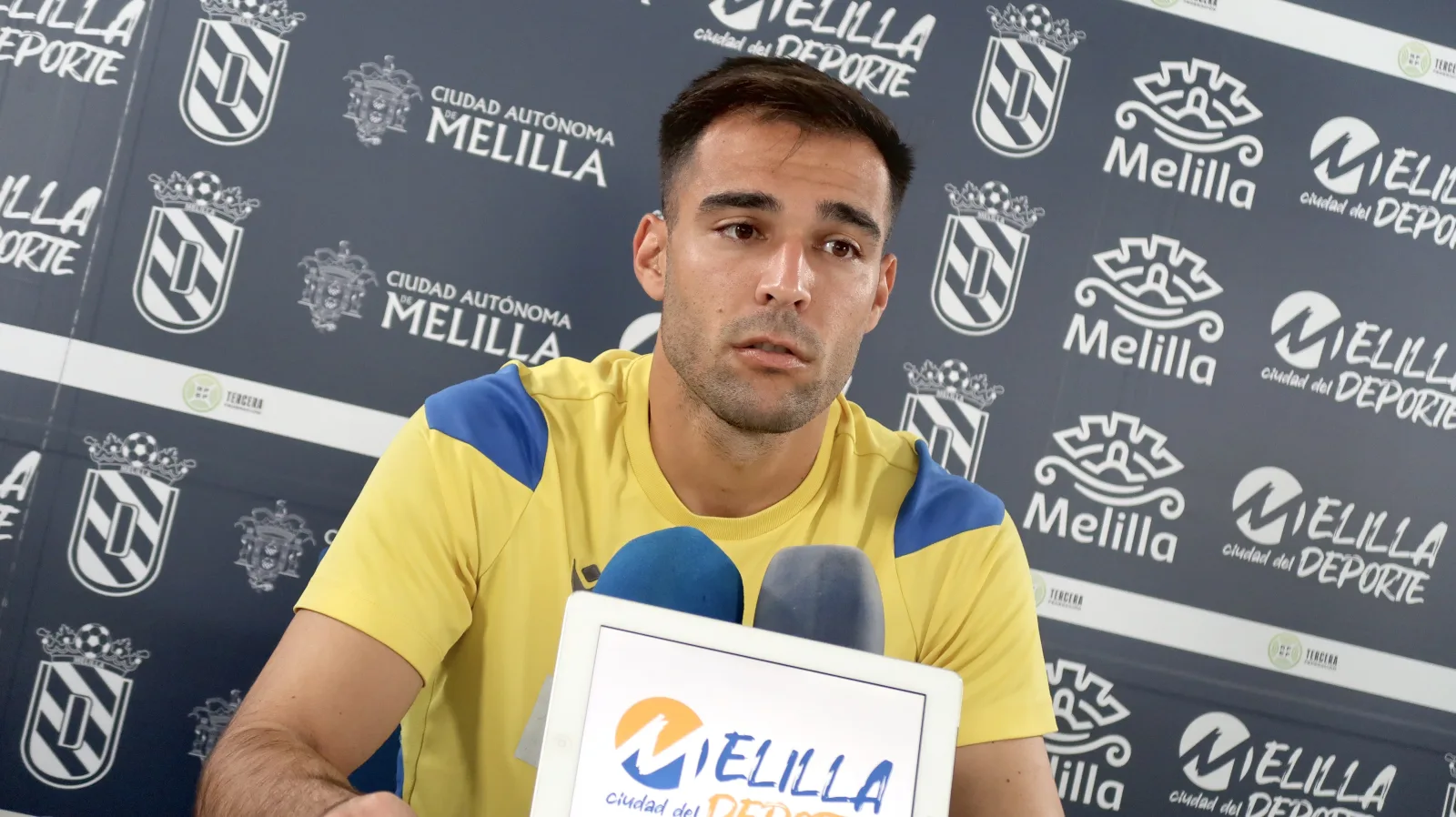 Un hombre joven con camiseta amarilla y azul habla ante micrófonos frente a un panel con escudos y el texto Melilla.