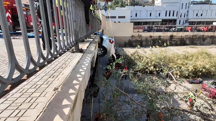 Un coche volcado cuelga del borde de un puente mientras bomberos con casco trabajan junto a él y bajo la estructura.