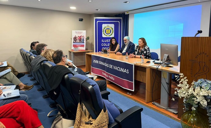 Personas sentadas en sillas azules frente a una mesa con tres ponentes y una pantalla, en una sala de conferencias.